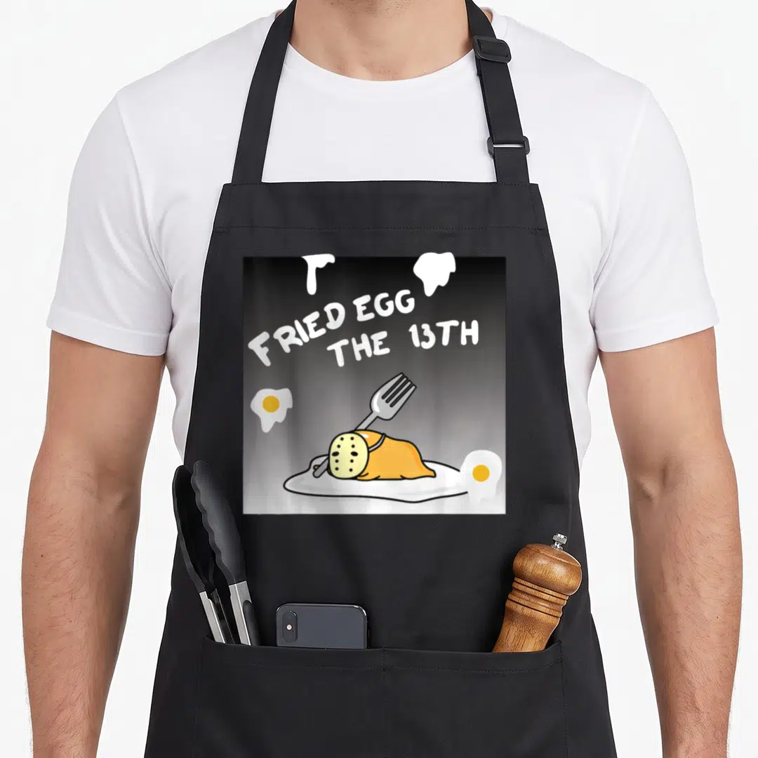 Fried egg apron - Cartoon masked fried egg stabbed by fork with FRIED EGG THE 13TH text — shown on a black cotton canvas apron against a white background