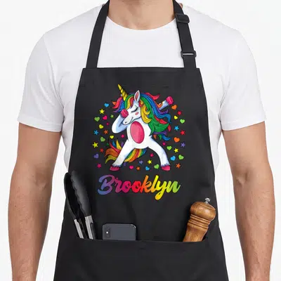 Brooklyn rainbow apron - White dabbing unicorn with rainbow mane surrounded by hearts and stars above rainbow text — shown on a black cotton canvas apron against a white background
