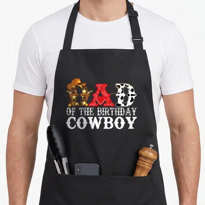 1st cowboy apron - DAD OF THE BIRTHDAY COWBOY text with western hat bandana and cow print letters — shown on a black cotton canvas apron against a white background
