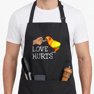 Hurt bird apron - Cartoon orange parrot biting finger with text LOVE HURTS — shown on a black cotton canvas apron against a white background