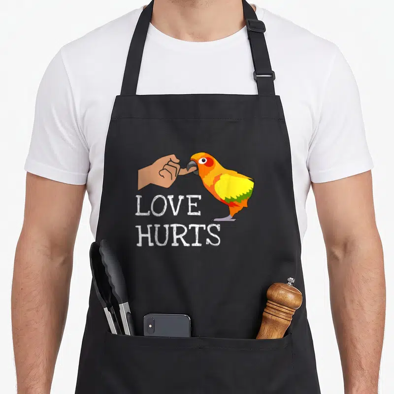 Hurt bird apron - Cartoon orange parrot biting finger with text LOVE HURTS — shown on a black cotton canvas apron against a white background