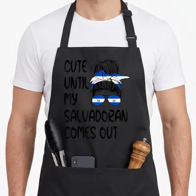 Come salvadoran apron - Stacked black text beside cartoon woman with Salvadoran flag headscarf and sunglasses in blue — shown on a black cotton canvas apron against a white background