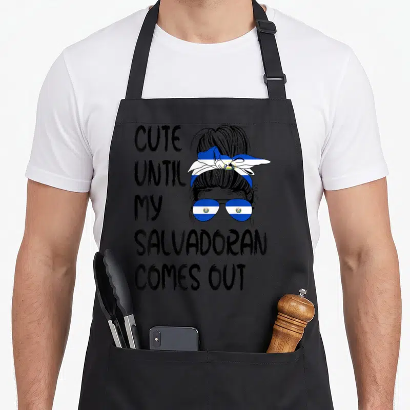 Come salvadoran apron - Stacked black text beside cartoon woman with Salvadoran flag headscarf and sunglasses in blue — shown on a black cotton canvas apron against a white background