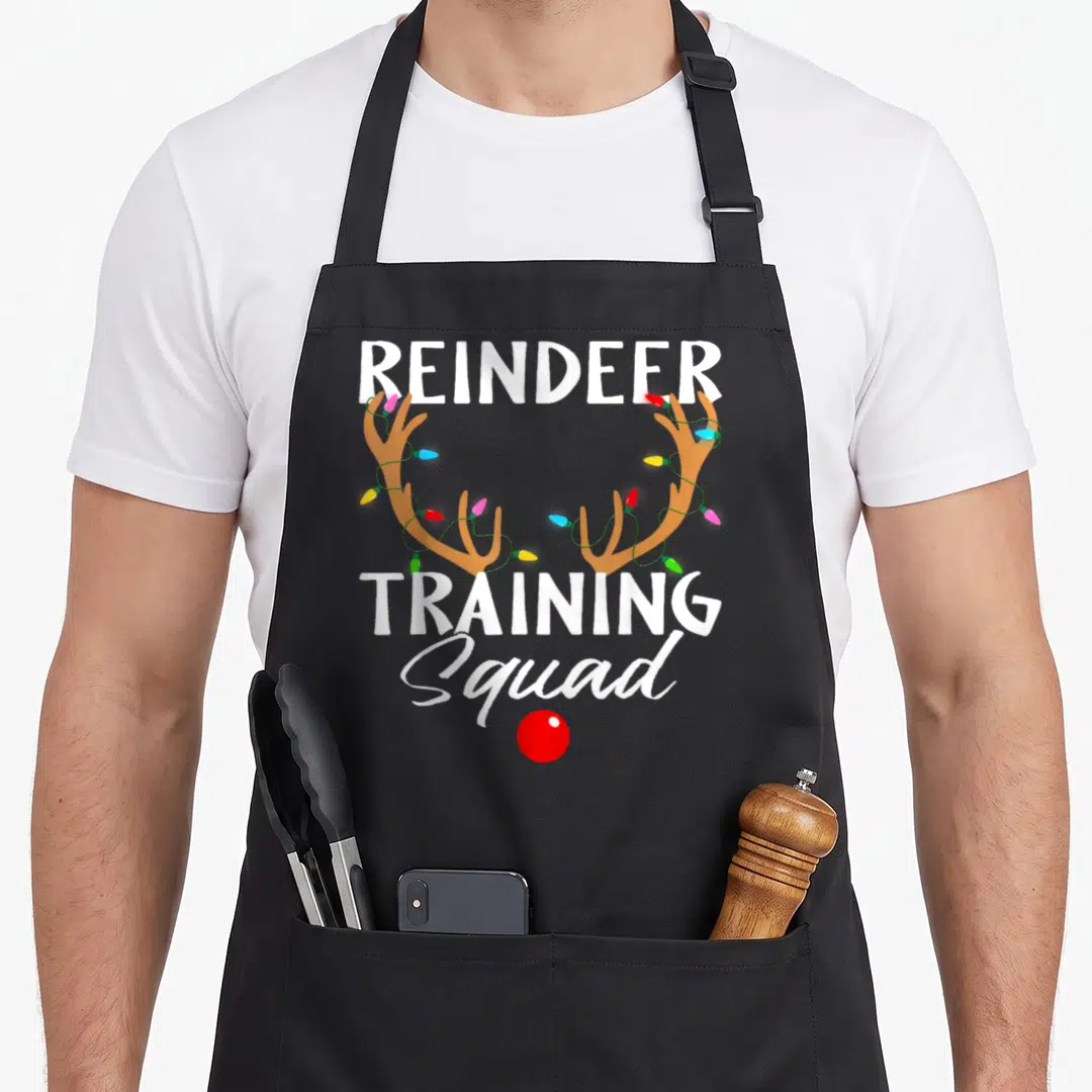 Running reindeer christmas apron - Cartoon antlers wrapped in lights with text REINDEER TRAINING Squad and red nose — shown on a black cotton canvas apron against a white background