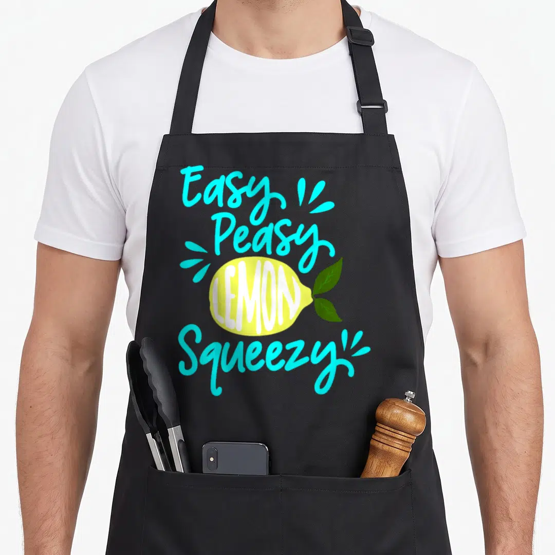 Peasy lemonade apron - Easy Peasy Lemon Squeezy text around yellow lemon with green leaves in cyan and white — shown on a black cotton canvas apron against a white background