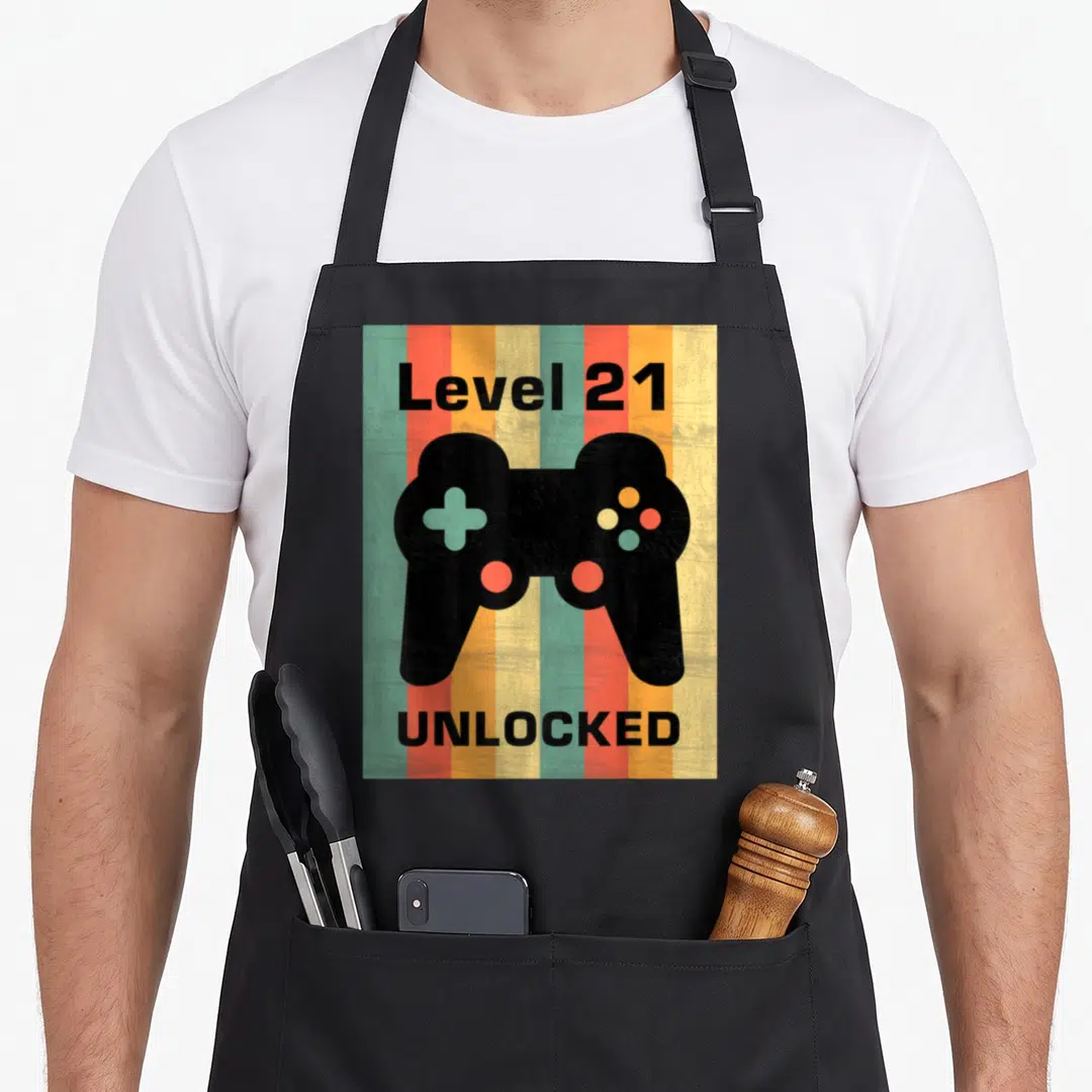 Gamepad birthday apron - Black game controller between Level 21 and UNLOCKED text on colored vertical stripes — shown on a black cotton canvas apron against a white background