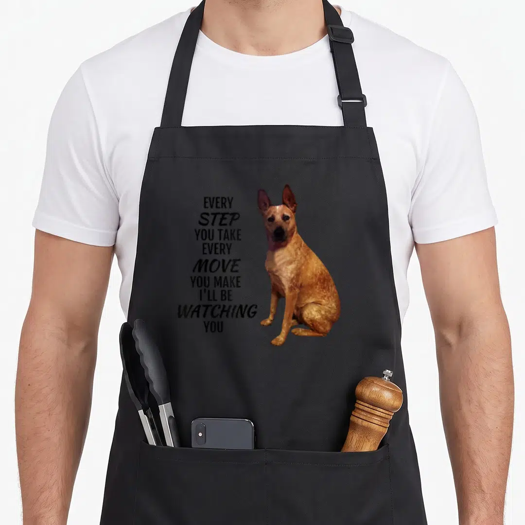 Step heeler apron - Sitting brown dog photo beside vertical black text reading Every Step You Take I'll Be Watching You — shown on a black cotton canvas apron against a white background