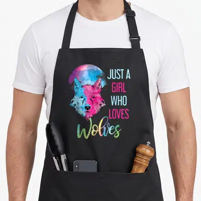 Moonlight predator apron - Cartoon wolf head in pink and blue with stacked text JUST A GIRL WHO LOVES Wolves — shown on a black cotton canvas apron against a white background