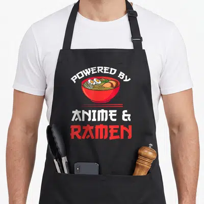 Merchandise anime apron - Red bowl with noodles and eggs under arched white text and stacked ANIME & RAMEN lettering — shown on a black cotton canvas apron against a white background