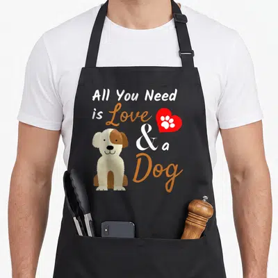 Funny present dog apron - Cartoon dog sitting beside stacked text reading All You Need is Love and a Dog with a red — shown on a black cotton canvas apron against a white background