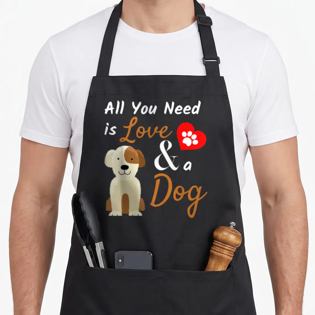 Funny present dog apron - Cartoon dog sitting beside stacked text reading All You Need is Love and a Dog with a red — shown on a black cotton canvas apron against a white background