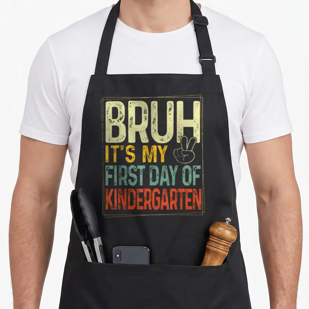 Bruh kindergarten back apron - Stacked distressed text BRUH IT'S MY FIRST DAY OF KINDERGARTEN with peace sign in beige — shown on a black cotton canvas apron against a white background