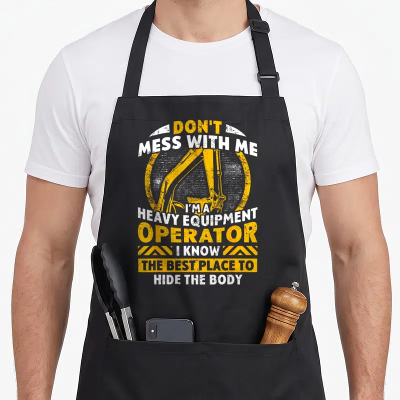 Equipment heavy apron - Excavator arm in yellow circle with sarcastic operator text and hazard stripes — shown on a black cotton canvas apron against a white background