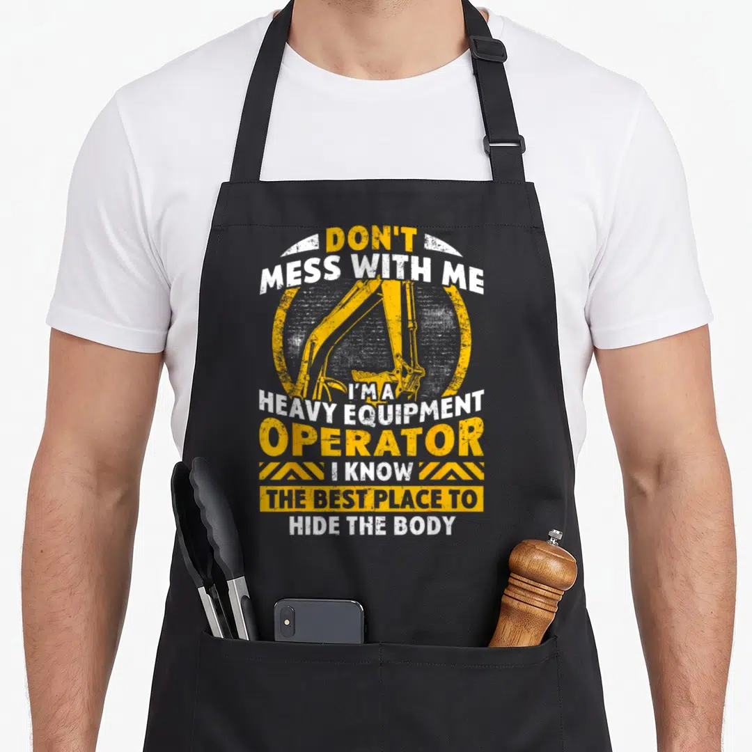 Equipment heavy apron - Excavator arm in yellow circle with sarcastic operator text and hazard stripes — shown on a black cotton canvas apron against a white background