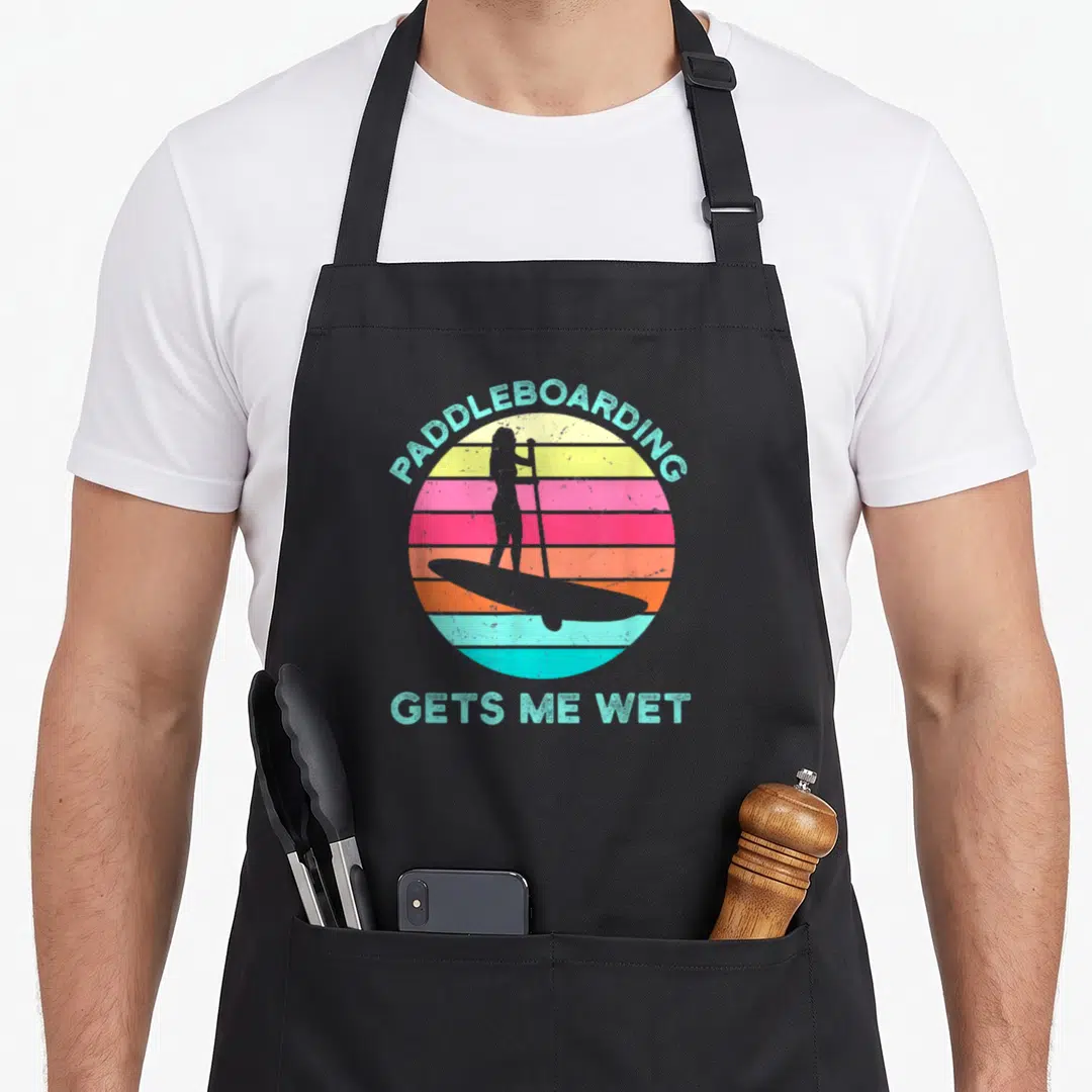 Paddleboarding wet aprons - Retro striped sun with paddleboarder silhouette and teal text PADDLEBOARDING GETS ME WET — shown on a black cotton canvas apron against a white background