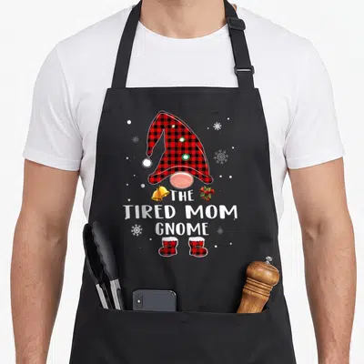 Tired buffalo plaid christmas apron - Cartoon gnome in red plaid hat and boots with text THE TIRED MOM GNOME flanked — shown on a black cotton canvas apron against a white background