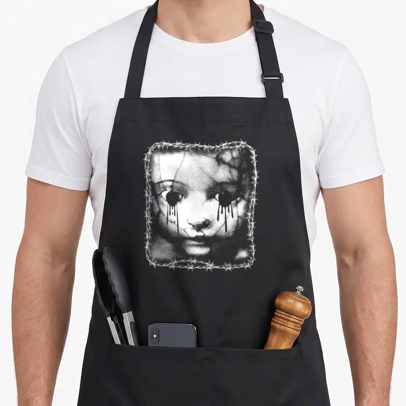 Wire goth apron - Distressed white face with dripping eyes inside barbed wire square frame — shown on a black cotton canvas apron against a white background
