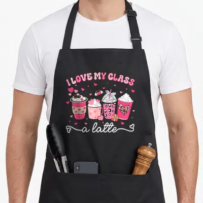 Valentine teacher apron - Four pink coffee cups with hearts and text I Love My Class A Latte — shown on a black cotton canvas apron against a white background