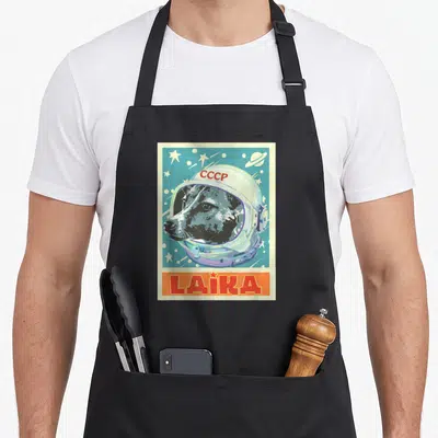 Soviet dog apron - Dog in space helmet with CCCP text and yellow LAIKA banner on blue starry background — shown on a black cotton canvas apron against a white background