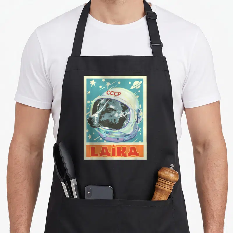Soviet dog apron - Dog in space helmet with CCCP text and yellow LAIKA banner on blue starry background — shown on a black cotton canvas apron against a white background