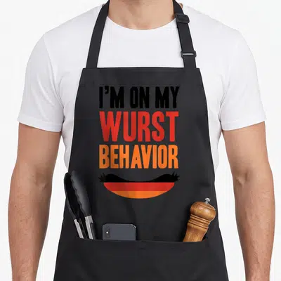 Wurst german oktoberfest aprons - Stacked black red and orange text reading I'M ON MY WURST BEHAVIOR above a German — shown on a black cotton canvas apron against a white background