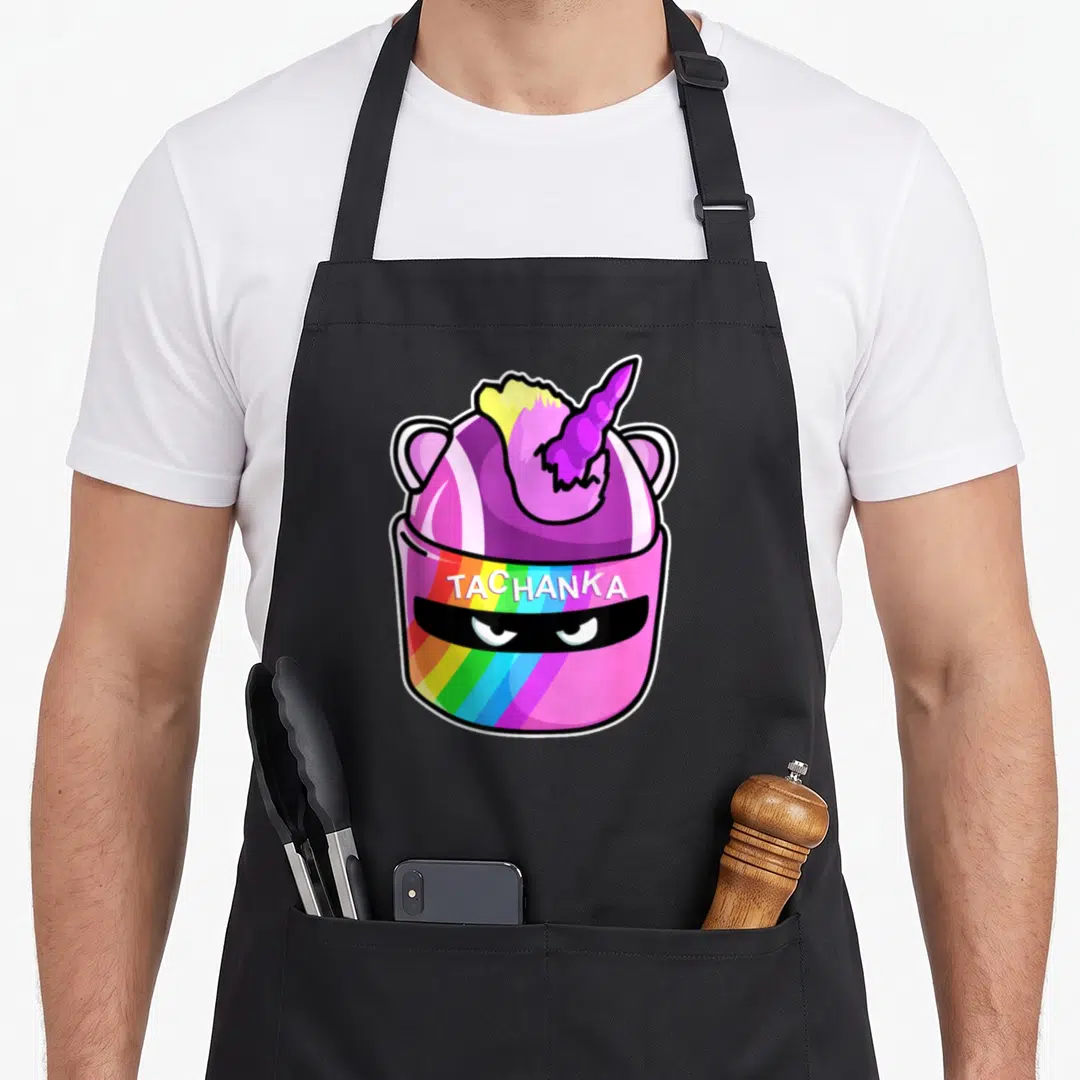 Komodo cartoon apron - Pink helmet with unicorn horn, rainbow stripes, and TACHANKA text — shown on a black cotton canvas apron against a white background