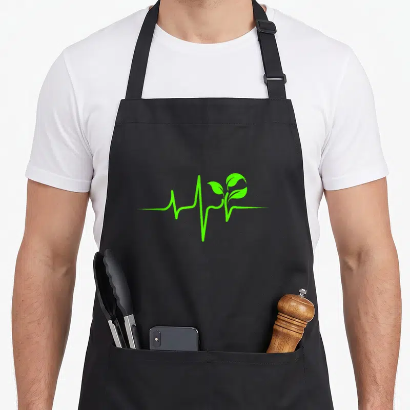 Whole plant based apron - Green heartbeat line with two leaf sprout on black bib — shown on a black cotton canvas apron against a white background