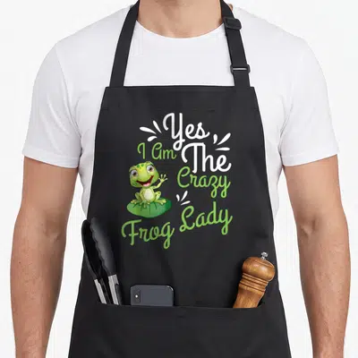 Cute toad frog aprons - Green cartoon frog on lily pad with Yes I Am The Crazy Frog Lady text — shown on a black cotton canvas apron against a white background
