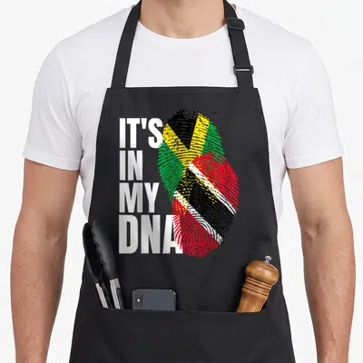 Trinidadian heritage aprons - IT'S IN MY DNA text beside two overlapping fingerprints filled with Jamaican flag colors — shown on a black cotton canvas apron against a white background