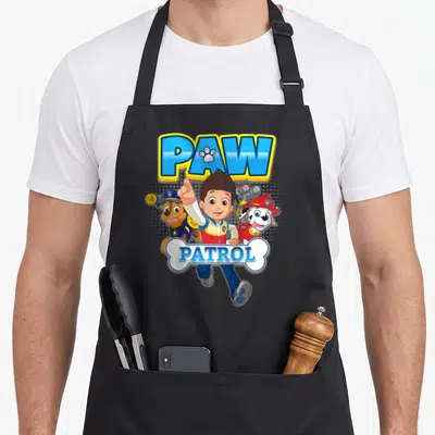 Ryder pup novelty apron - Ryder pointing up between Chase and Marshall under PAW PATROL bone logo — shown on a black cotton canvas apron against a white background