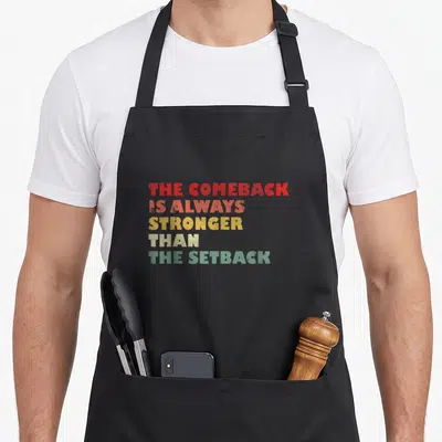 Alway stronger comeback apron - Stacked vintage text reading THE COMEBACK IS ALWAYS STRONGER THAN THE SETBACK in red — shown on a black cotton canvas apron against a white background