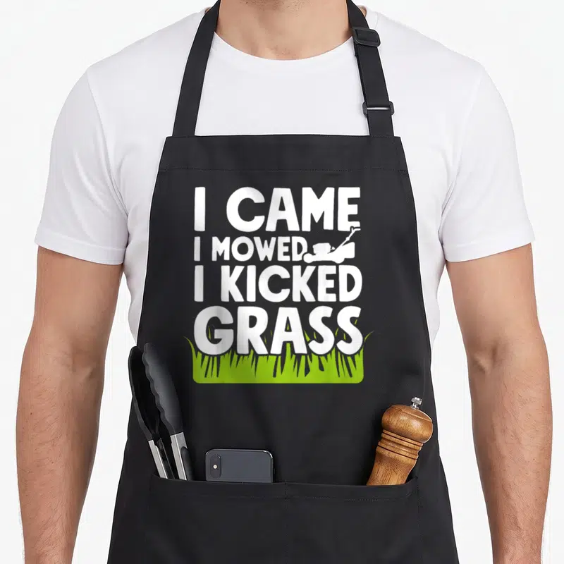 Came yard apron - White stacked text I Came I Mowed I Kicked Grass with small mower silhouette and green grass patch — shown on a black cotton canvas apron against a white background