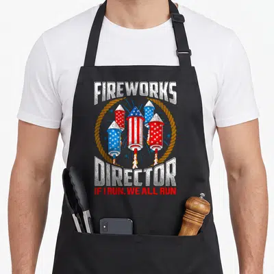 Director 4th of july apron - Distressed fireworks director graphic with four patriotic rockets and safety warning text — shown on a black cotton canvas apron against a white background