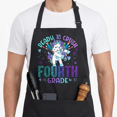Fourth unicorn apron - Dabbing unicorn with fanned pencils and brushes, arched Ready to Crush Fourth Grade text — shown on a black cotton canvas apron against a white background