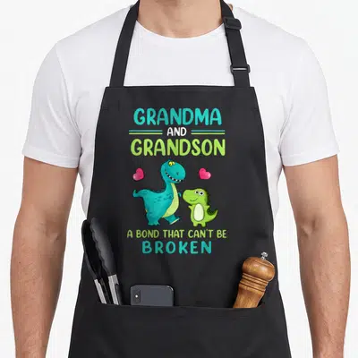 Broken grandma apron - Teal and lime dinosaurs with pink hearts and bond text — shown on a black cotton canvas apron against a white background