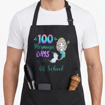 School mermaid apron - Cartoon mermaid with 100 Mermagic DAYS Of School text and three stars in turquoise and purple — shown on a black cotton canvas apron against a white background