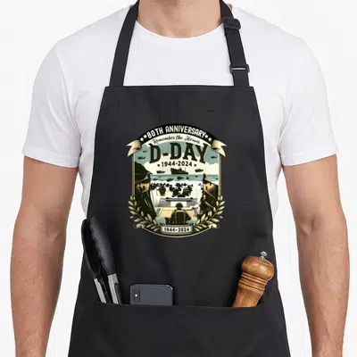 Remembrance military apron - Retro D-Day emblem showing soldiers landing on a beach with ships and 1944-2024 text — shown on a black cotton canvas apron against a white background