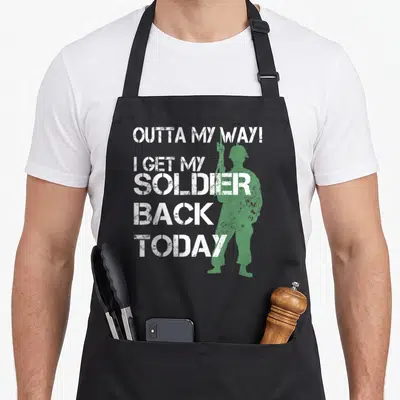Outta homecoming apron - White stencil text Outta My Way I Get My Soldier Back Today with green soldier silhouette — shown on a black cotton canvas apron against a white background