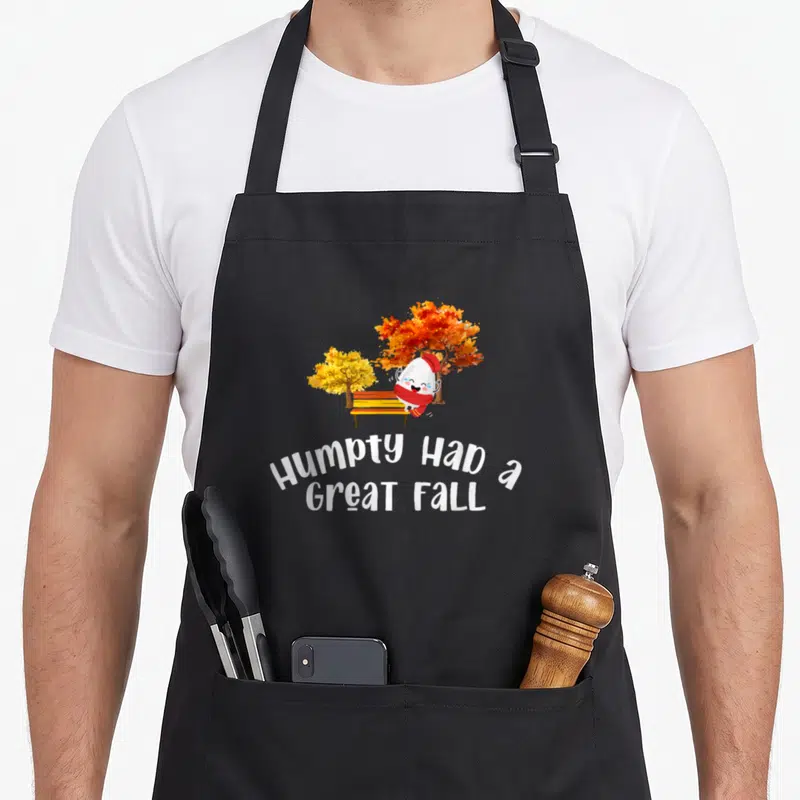 Cute humpty cute fall aprons - Cartoon egg on bench with two autumn trees and curved white text reading Humpty Had — shown on a black cotton canvas apron against a white background