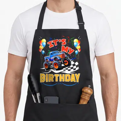 Monster car truck apron - Blue monster truck with IT'S MY BIRTHDAY text, balloons, and checkered flag in red yellow — shown on a black cotton canvas apron against a white background