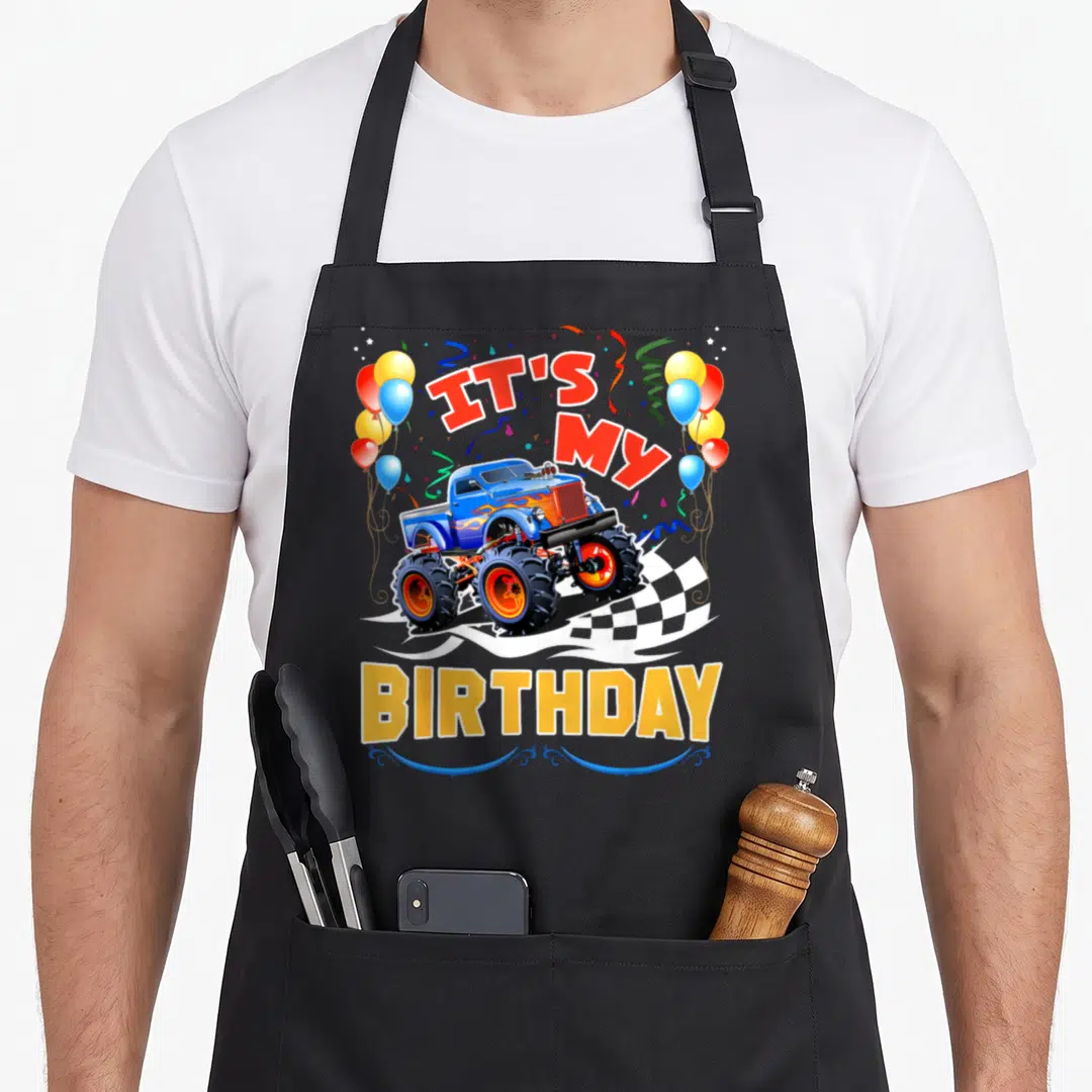 Monster car truck apron - Blue monster truck with IT'S MY BIRTHDAY text, balloons, and checkered flag in red yellow — shown on a black cotton canvas apron against a white background