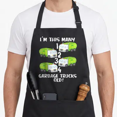 Counting truck apron - Four green cartoon garbage trucks numbered one to four with text I'M THIS MANY GARBAGE TRUCKS — shown on a black cotton canvas apron against a white background
