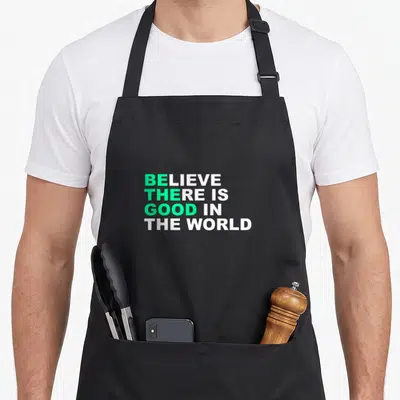 Believe positive vibes apron - Stacked green and white text reading BELIEVE THERE IS GOOD IN THE WORLD — shown on a black cotton canvas apron against a white background