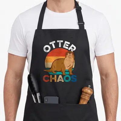 Punny animal apron - Retro sun with brown otter and OTTER CHAOS text in orange yellow blue white — shown on a black cotton canvas apron against a white background