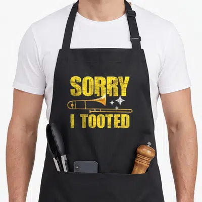 Tooted trombone apron - Yellow distressed trombone between SORRY and I TOOTED text with three white sparkles — shown on a black cotton canvas apron against a white background