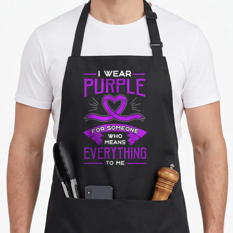 Hodgkin caregiver apron - Purple heart ribbon with vertical text I wear purple for someone who means everything to me — shown on a black cotton canvas apron against a white background