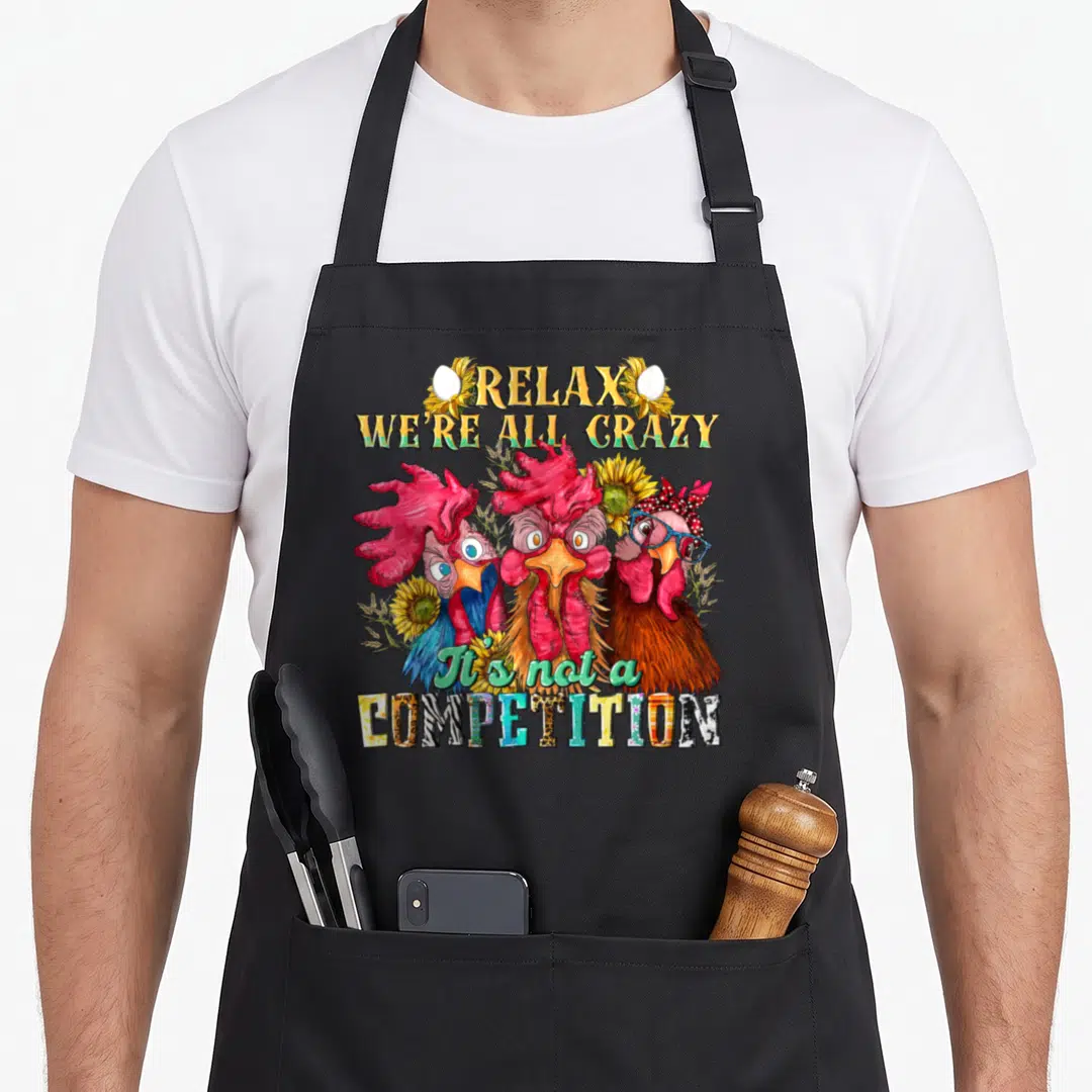 Relax chicken apron - Cartoon chickens with RELAX WE'RE ALL CRAZY text, sunflowers, eggs, and patterned COMPETITION — shown on a black cotton canvas apron against a white background