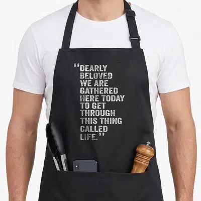Dearly today apron - White distressed text stack reading Dearly Beloved We Are Gathered Here Today To Get Through This — shown on a black cotton canvas apron against a white background
