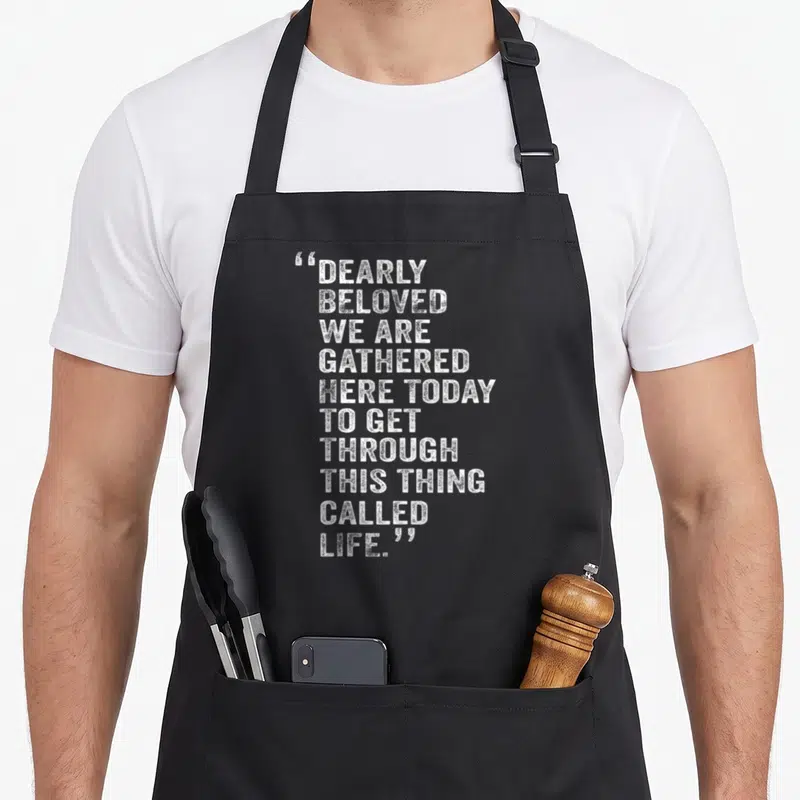 Dearly today apron - White distressed text stack reading Dearly Beloved We Are Gathered Here Today To Get Through This — shown on a black cotton canvas apron against a white background