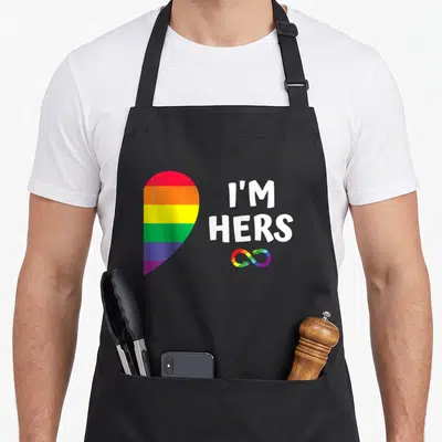 Pride she's in her apron - Rainbow heart next to white I'M HERS text and colored infinity symbol — shown on a black cotton canvas apron against a white background